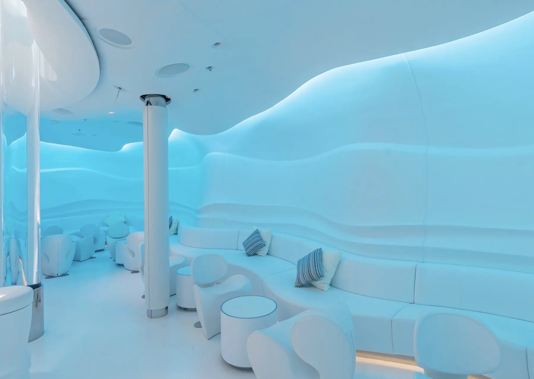 Blau beleuchtete Lounge der Spray Bar auf der AIDAperla mit geschwungenen weißen Sitzflächen