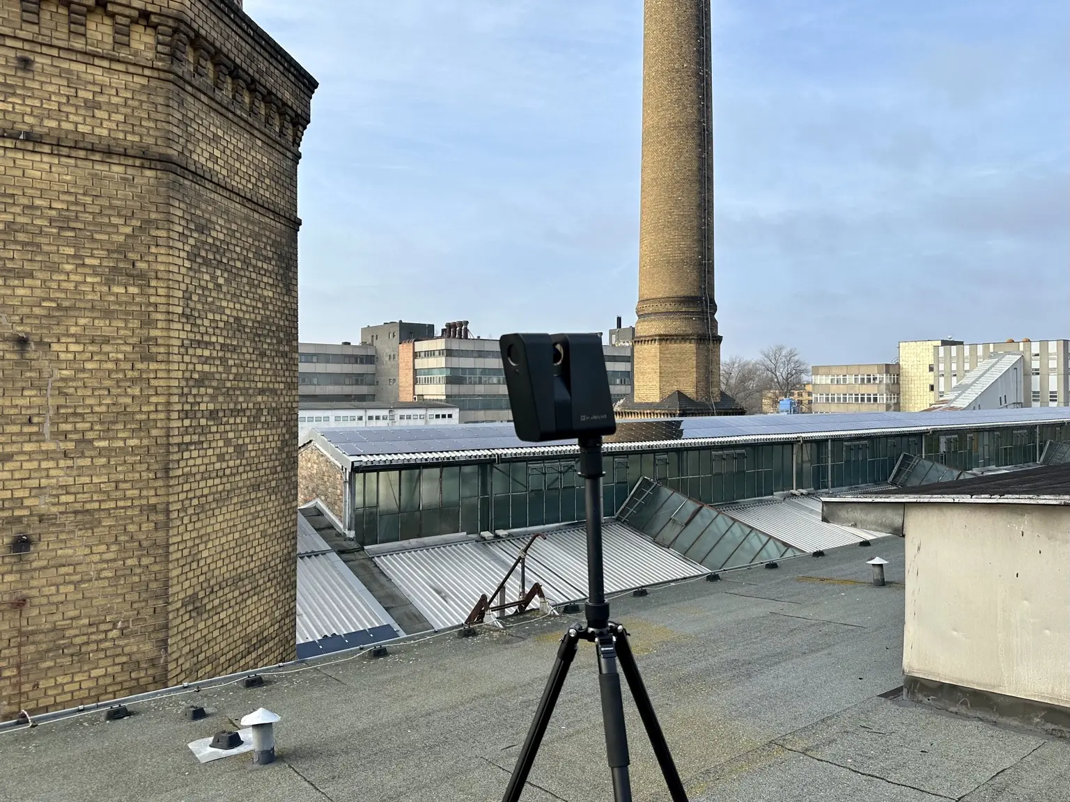 3D-Scanner auf einem Hallendach vor dem Schornstein und den Sheddächern der Rathenau-Hallen.
