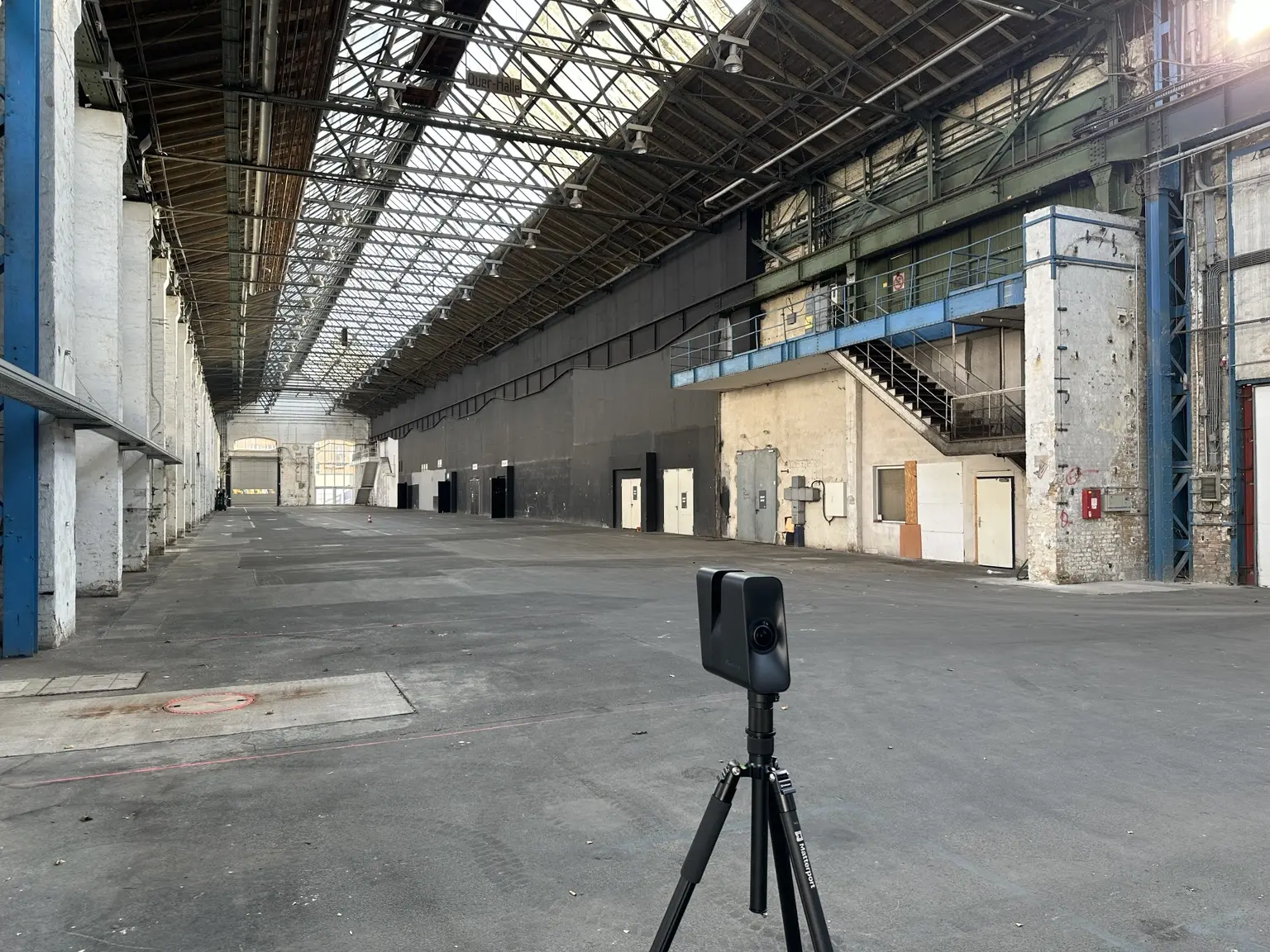 3D-Scanner vor einer langen Querhalle mit Stahltragwerk, Laufsteg und industrieller Bestandsstruktur.